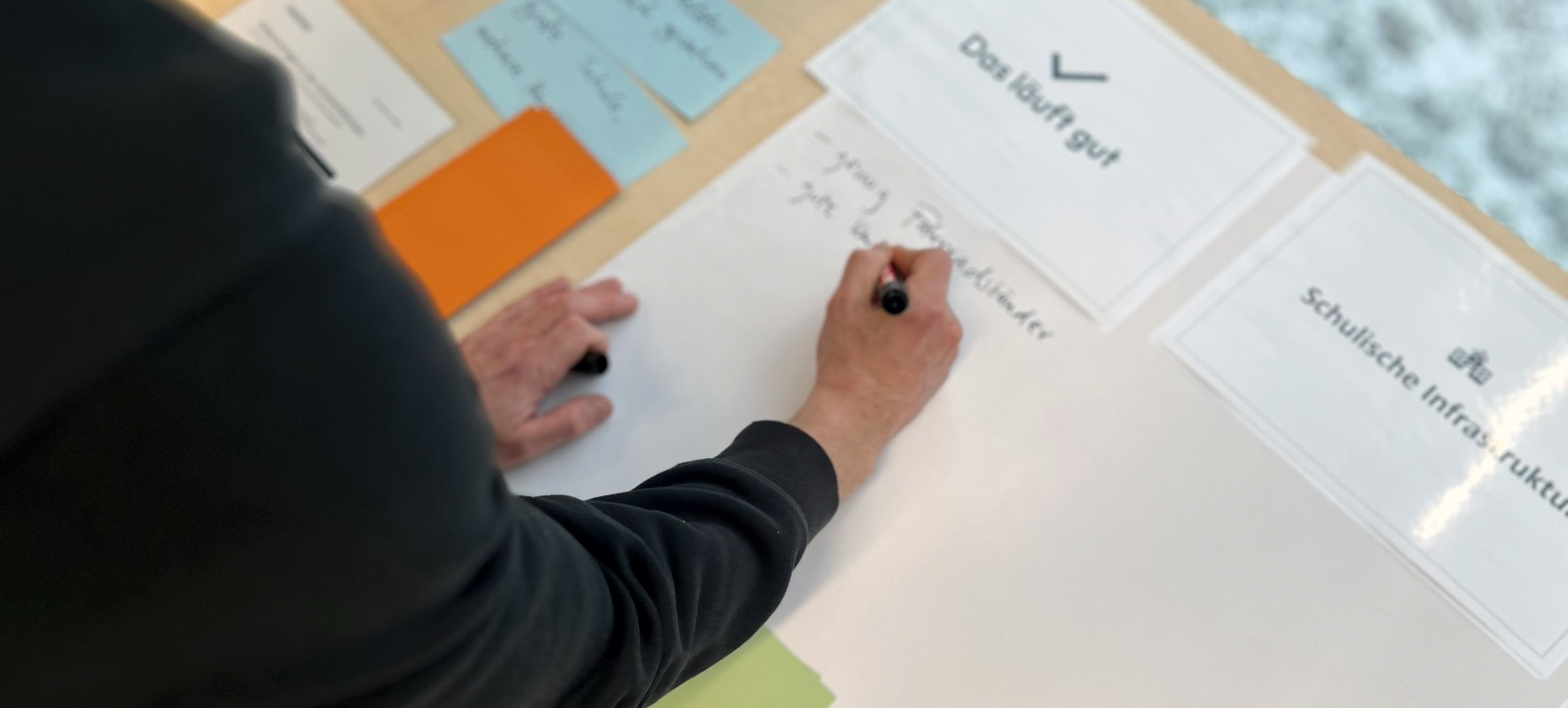 Eine Hand schreibt auf einem Papier, das auf einem Tisch liegt. Darüber liegen zwei laminierte Zettel, auf einem steht "Das läuft gut", auf dem anderen "Schulsche Infrastruktur"